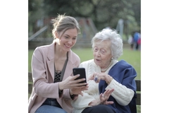 Old woman and younger woman sitting on a bench in the park, looking at a phone.